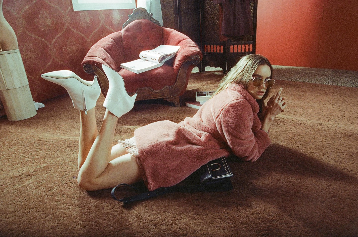 A woman in a pink coat and white boots lies on a brown carpet in a room with a red armchair and patterned wallpaper.