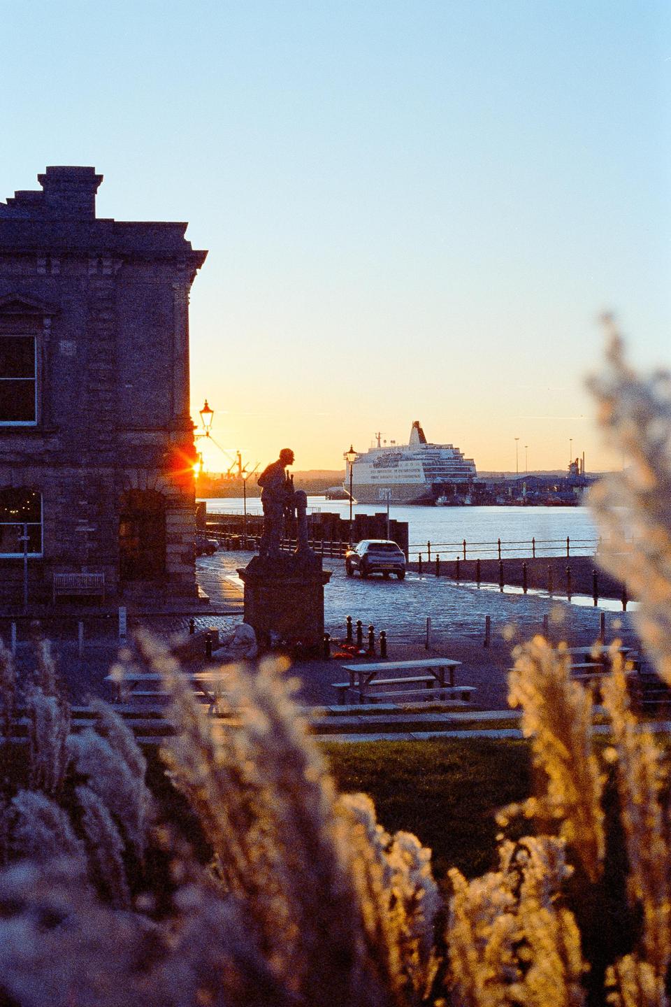 A statue of a man stands on a cobblestone street next to a body of water with a large ship in the distance at sunset.