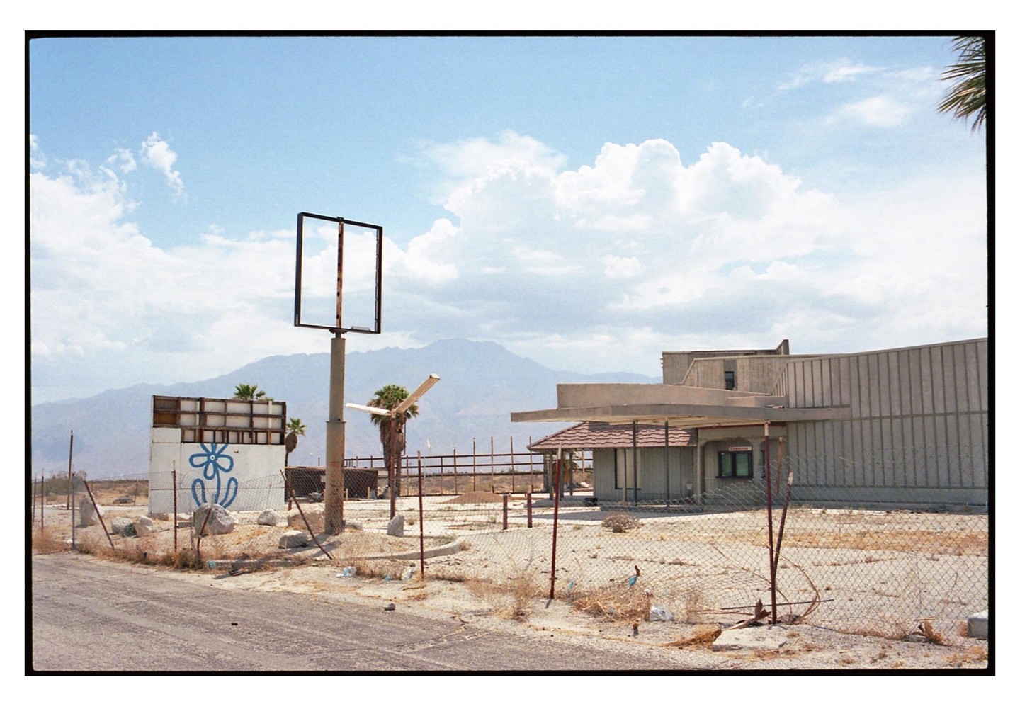 A deserted building with a large empty sign stands in a dry, dusty landscape with mountains in the background.