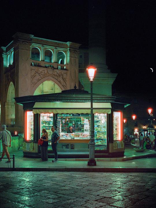 A couple stands in front of a brightly lit newsstand at night on a cobblestone street.