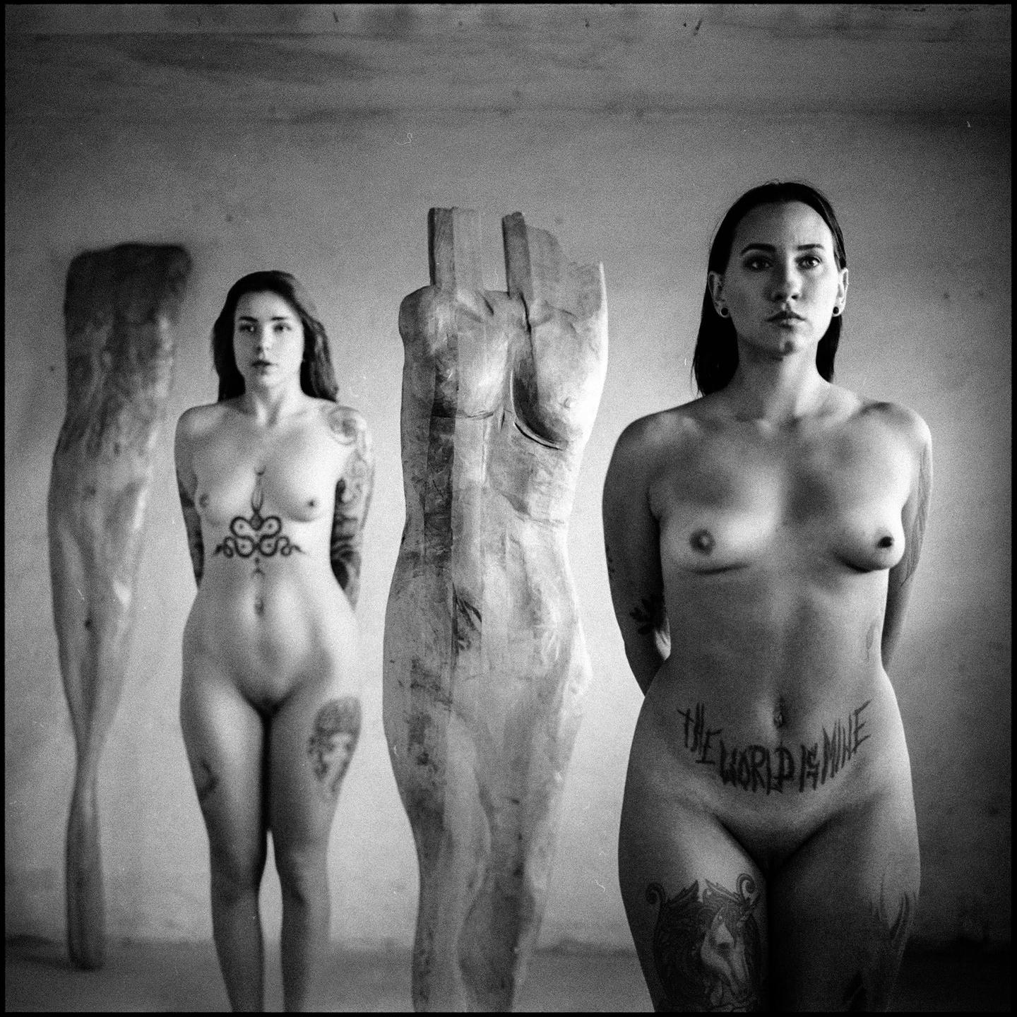 Two women with tattoos stand nude between three wooden sculptures of torsos.