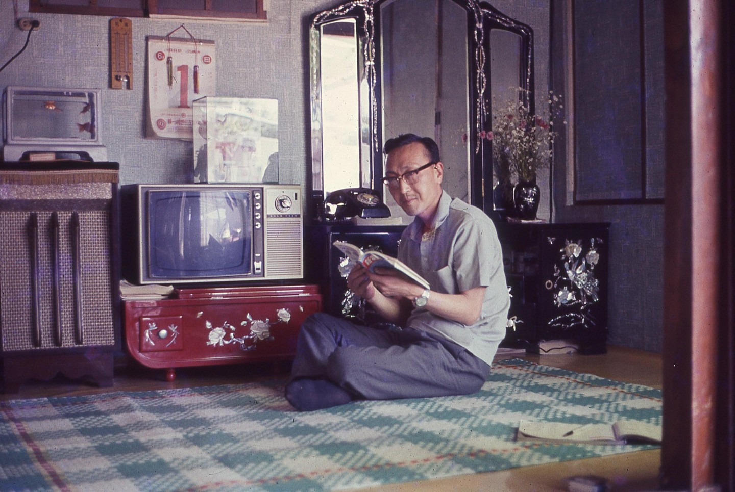 A man sits on a patterned rug, reading a book, with a television, a fish tank, and ornate furniture around him.