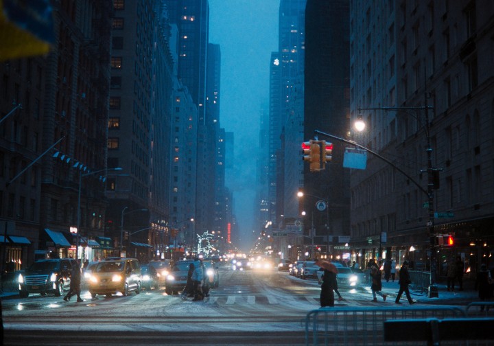 A snowy street in New York City is filled with cars and pedestrians at dusk, with tall buildings lining both sides of the road.