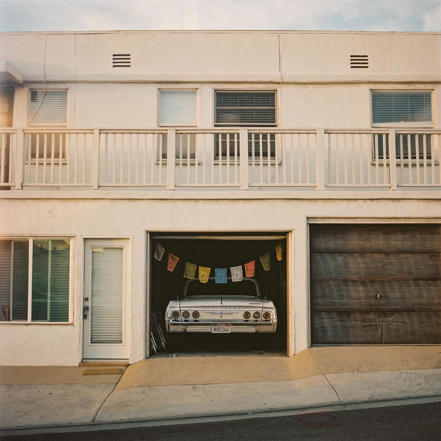 A white car is parked in a garage with prayer flags hanging above it, next to a closed wooden garage door and a white door.