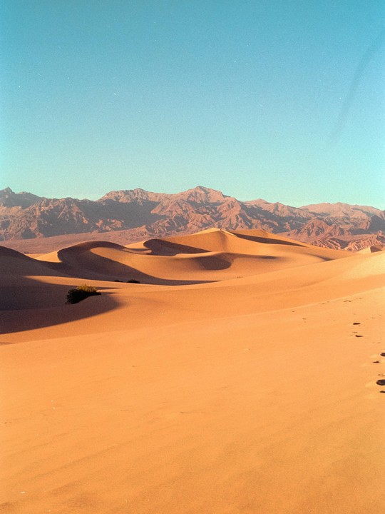 Sand dunes with shadows stretch towards mountains under a clear sky.