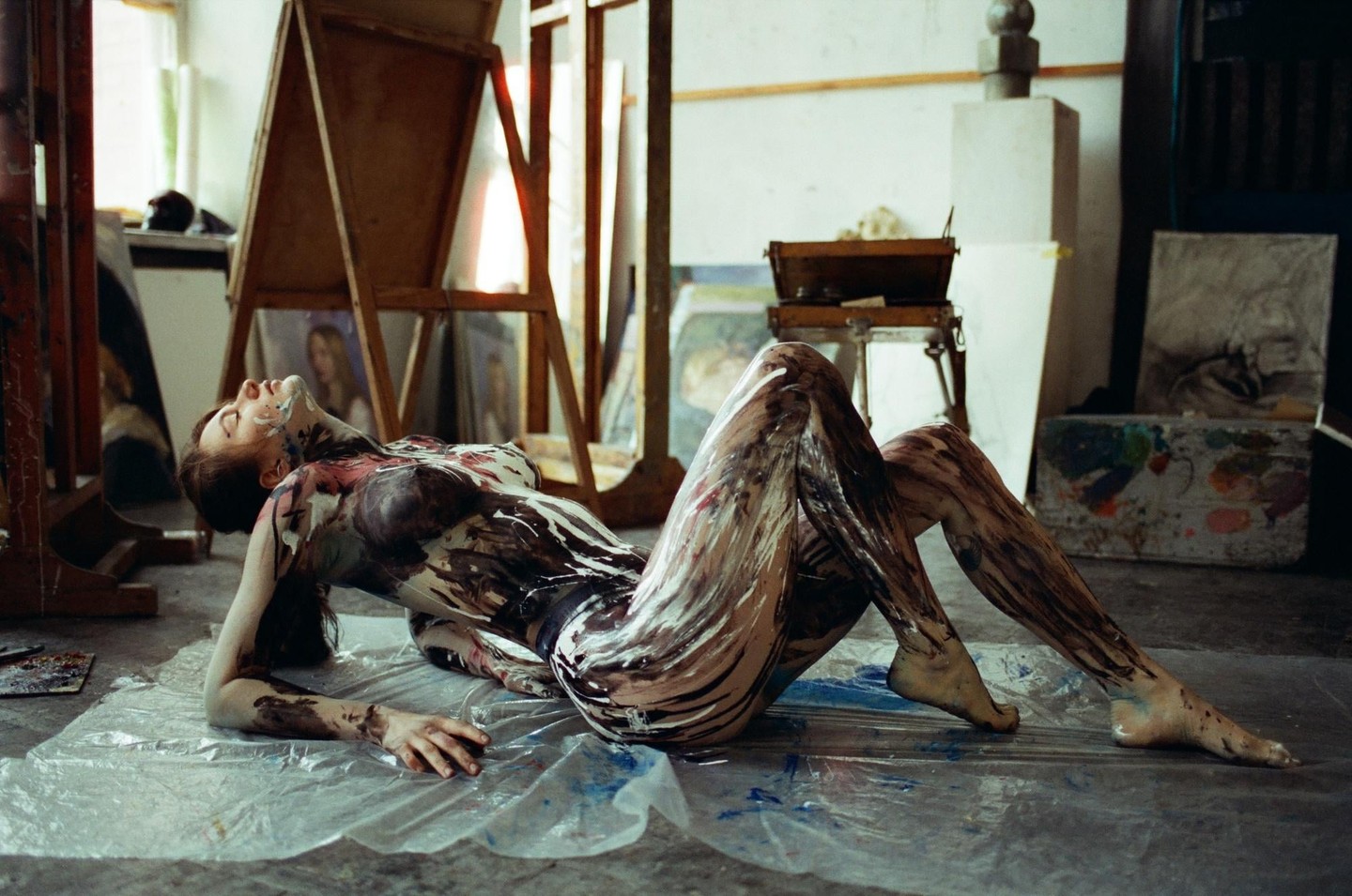 A woman covered in paint lies on a plastic sheet in an art studio.