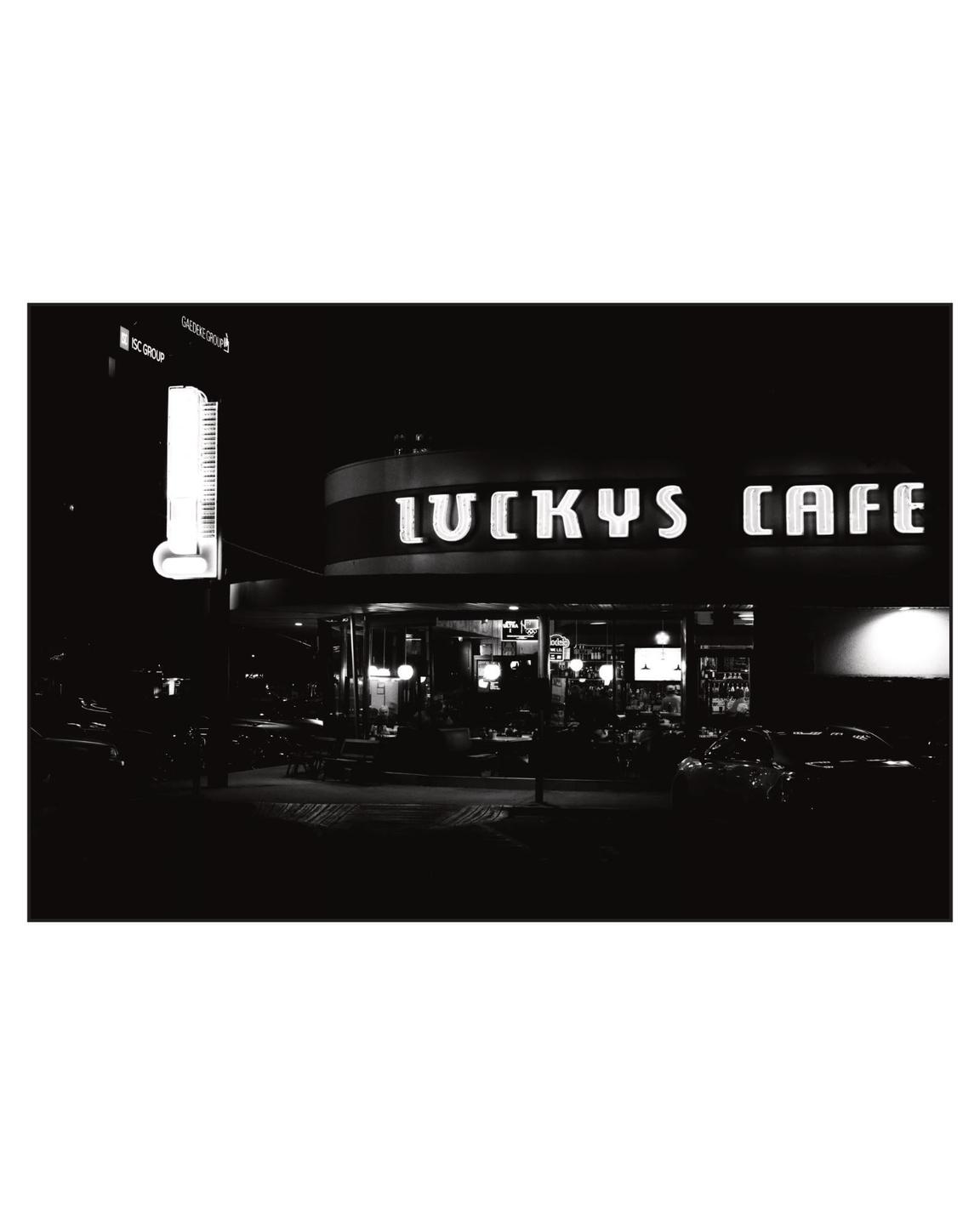 A black and white night street view shows the exterior of Lucky's Cafe with its neon sign illuminated.