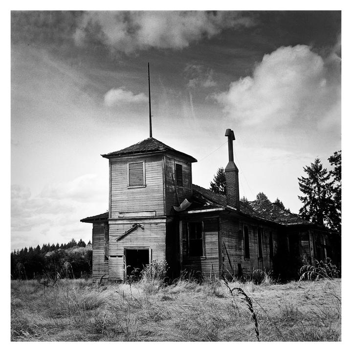An old, abandoned schoolhouse with a tower and chimney stands in a field of tall grass under a cloudy sky.