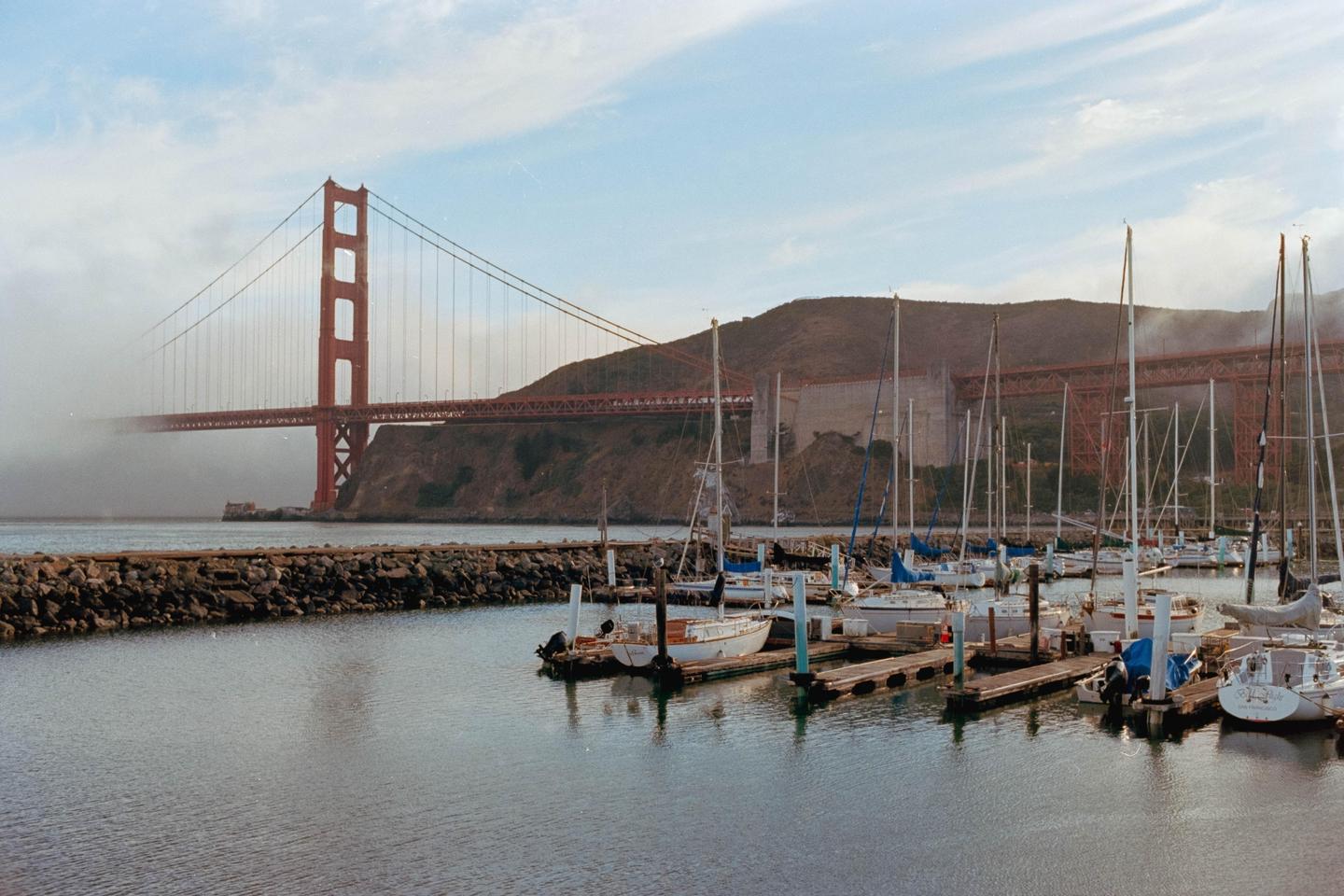 A marina with many sailboats is in the foreground, with the Golden Gate Bridge and a hazy mountain in the background.