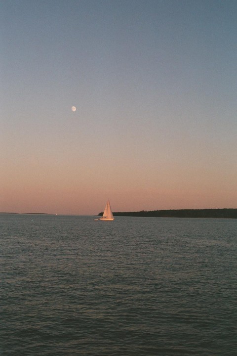 A sailboat is on the water with a moon in the sky and a treeline in the distance.