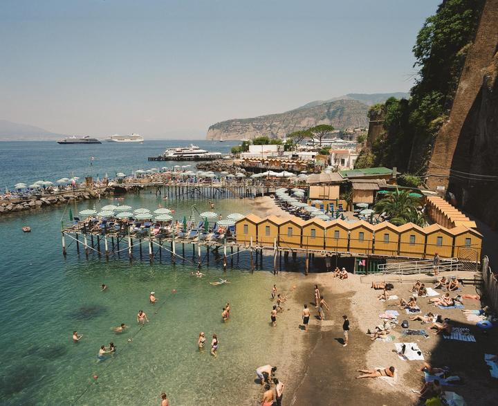 A crowded beach with yellow changing cabins and striped umbrellas is visible next to a calm sea with people swimming and large ships in the distance.
