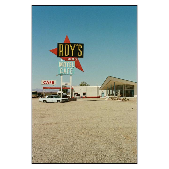 A large sign for Roy's Motel and Cafe stands in a gravel lot with a building and cars behind it.