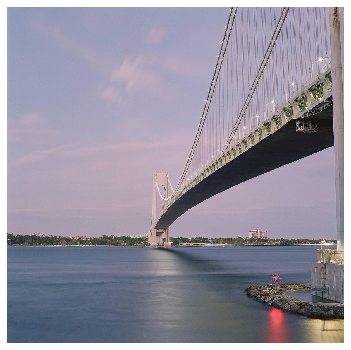 A long exposure captures the Verrazzano-Narrows Bridge illuminated at dusk, with its reflection visible on the water.