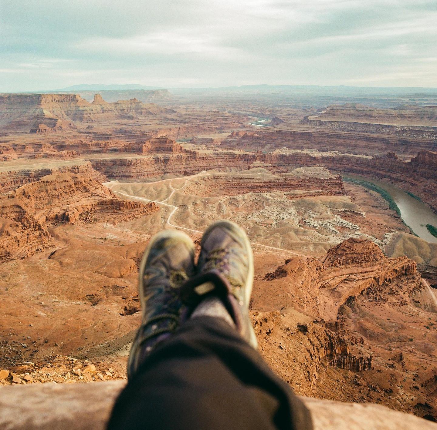 A person's feet and legs are visible in the foreground, overlooking a vast canyon landscape with a river winding through it.