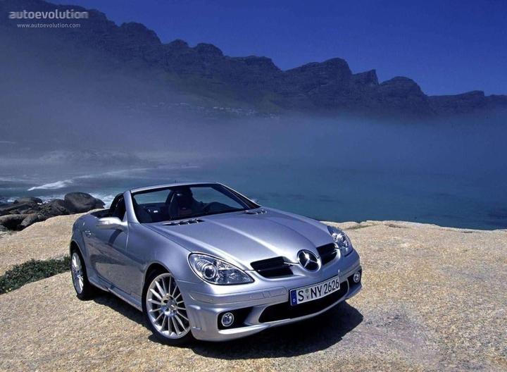 A silver convertible car is parked on a rocky cliff overlooking a body of water with mountains in the background.