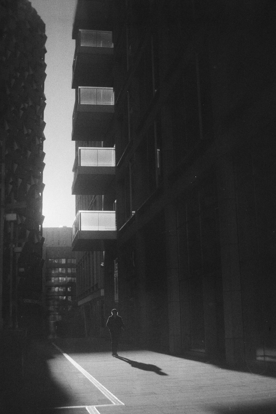 A man walks down a street between tall buildings, casting a long shadow.