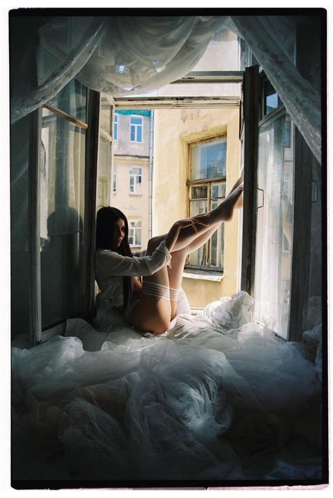 A woman in lingerie sits on a window sill with her legs raised, looking out at a building across the street.