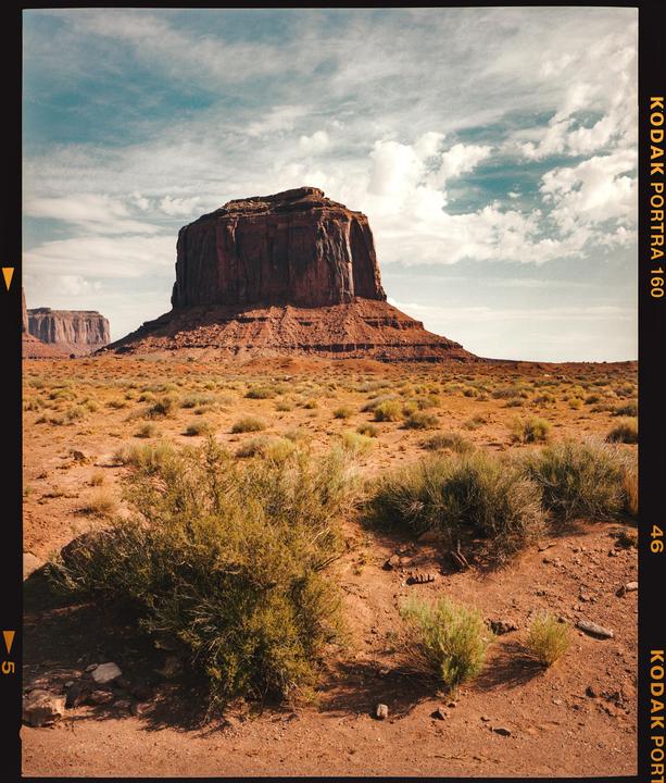 A large red rock formation stands in a desert landscape under a cloudy sky.