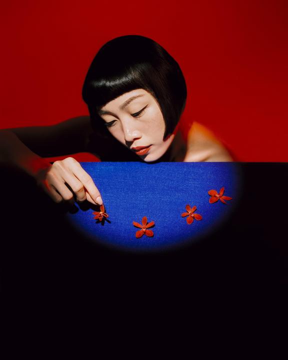A woman with short dark hair and red lipstick is looking down at small red flowers on a blue surface, with a red background.