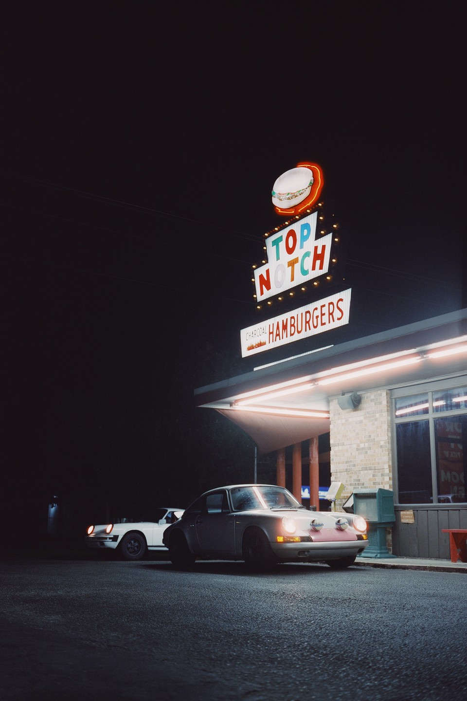 Two cars are parked at night in front of a hamburger restaurant with a neon sign.