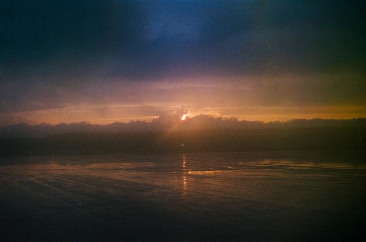 The sun sets behind a cloud bank over a body of water, casting reflections on the surface.