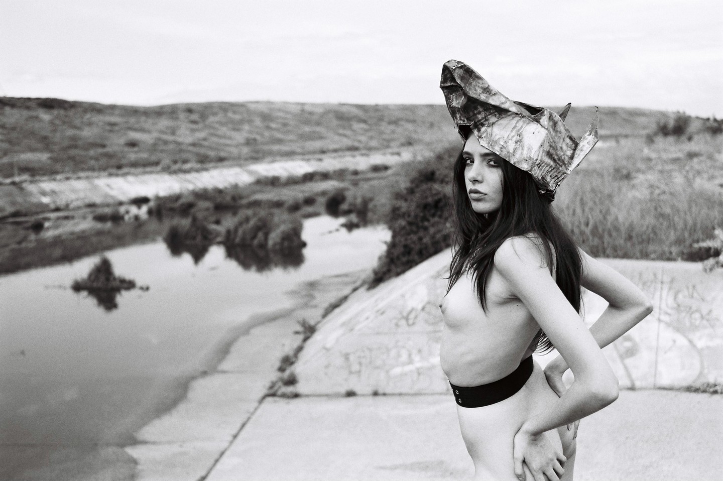 A woman wearing a metal hat stands next to a river with a concrete bank.