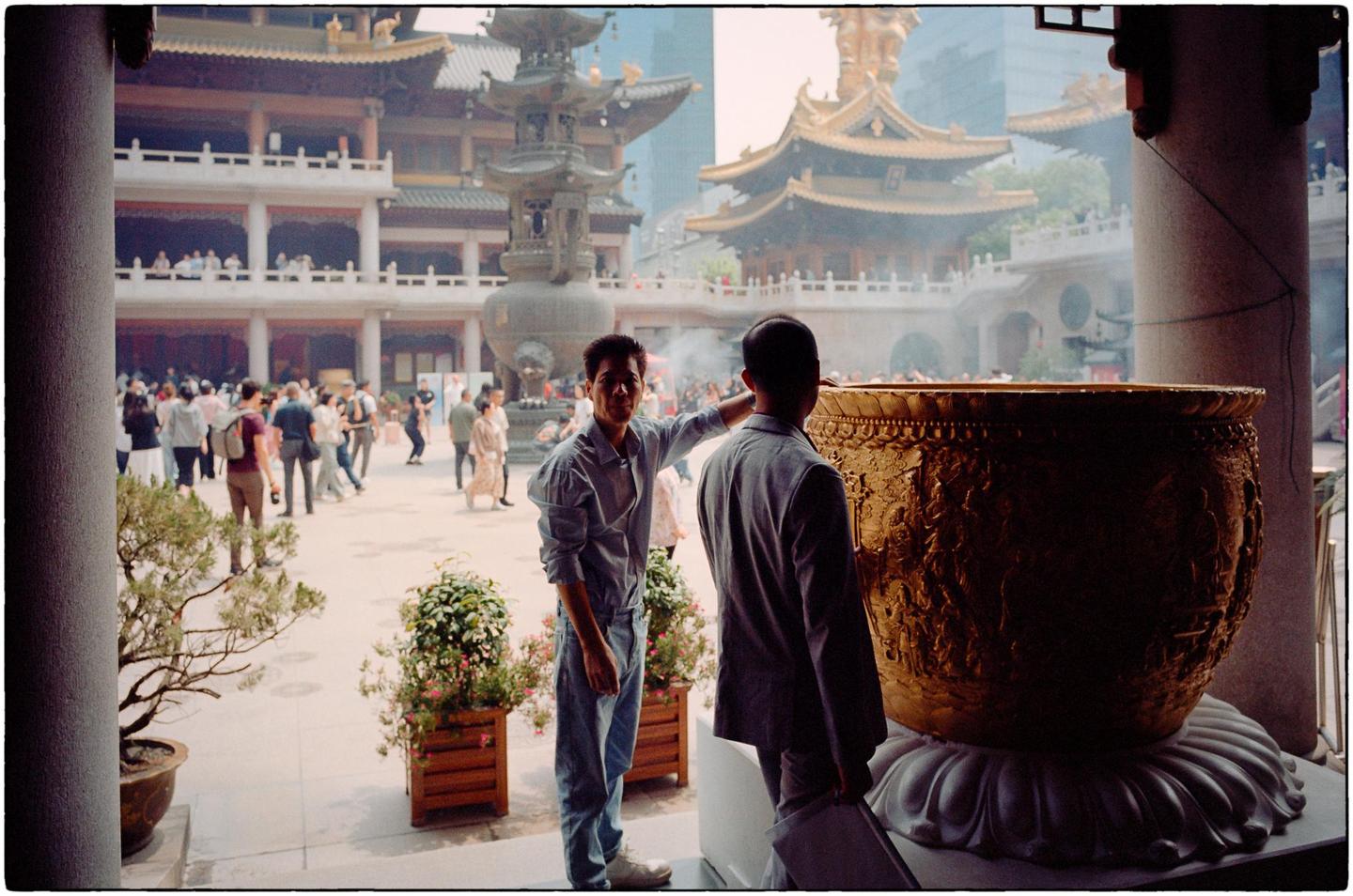 A man in a blue shirt smiles at the camera while another man in a suit stands with his back to the camera next to a large golden urn in a temple courtyard.