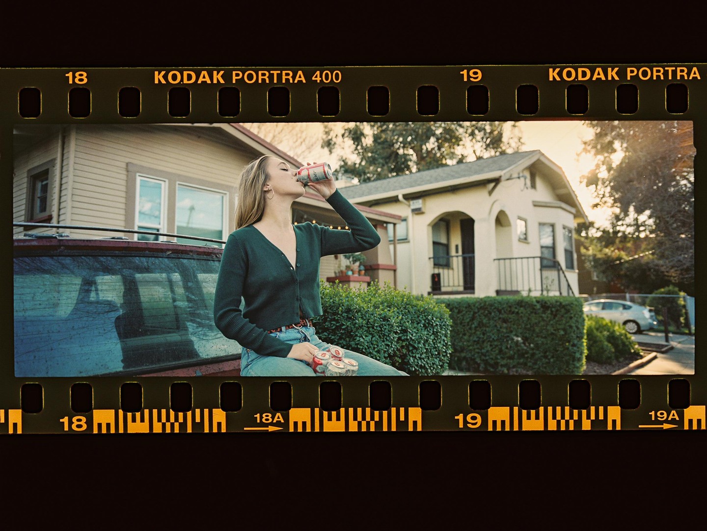 A woman sits on the back of a car and drinks from a can in front of a house.