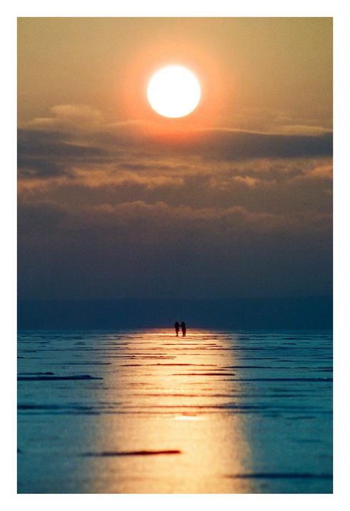 A couple stands on a frozen lake with the setting sun reflecting on the ice.