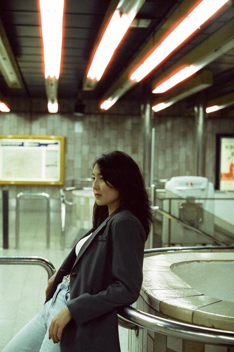 A woman in a dark blazer and light jeans leans against a railing in a subway station.