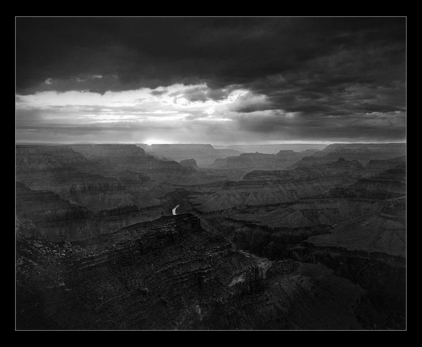A river winds through a vast canyon under a dramatic, cloudy sky with visible sun rays.