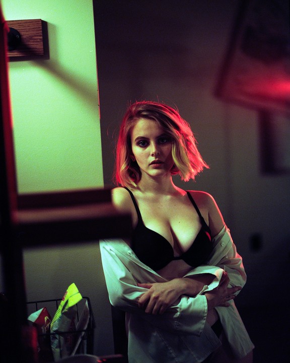 A woman in a bra and open shirt stands in a room with red and green lighting.