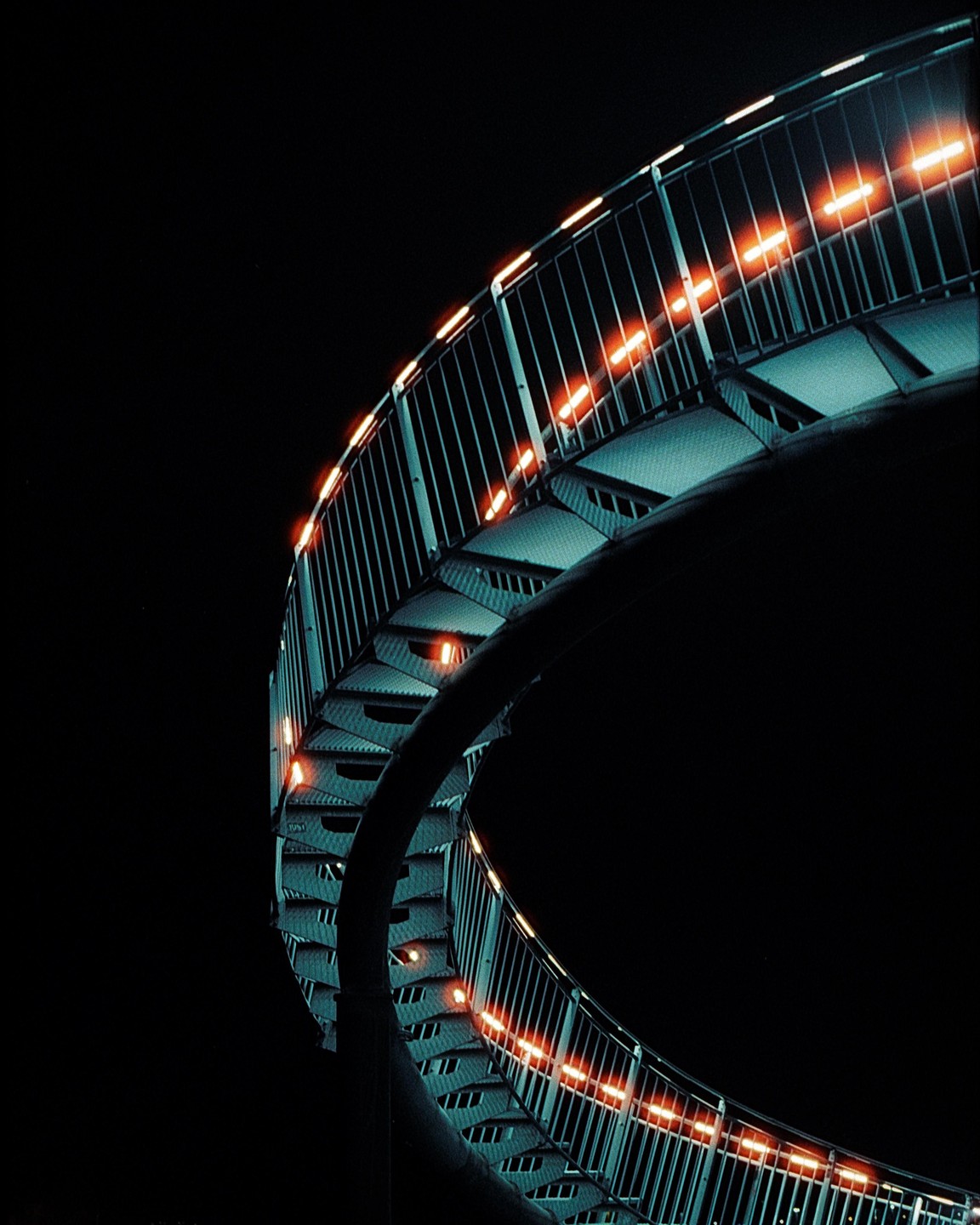 A spiral staircase with orange lights on the railing ascends into a dark sky.
