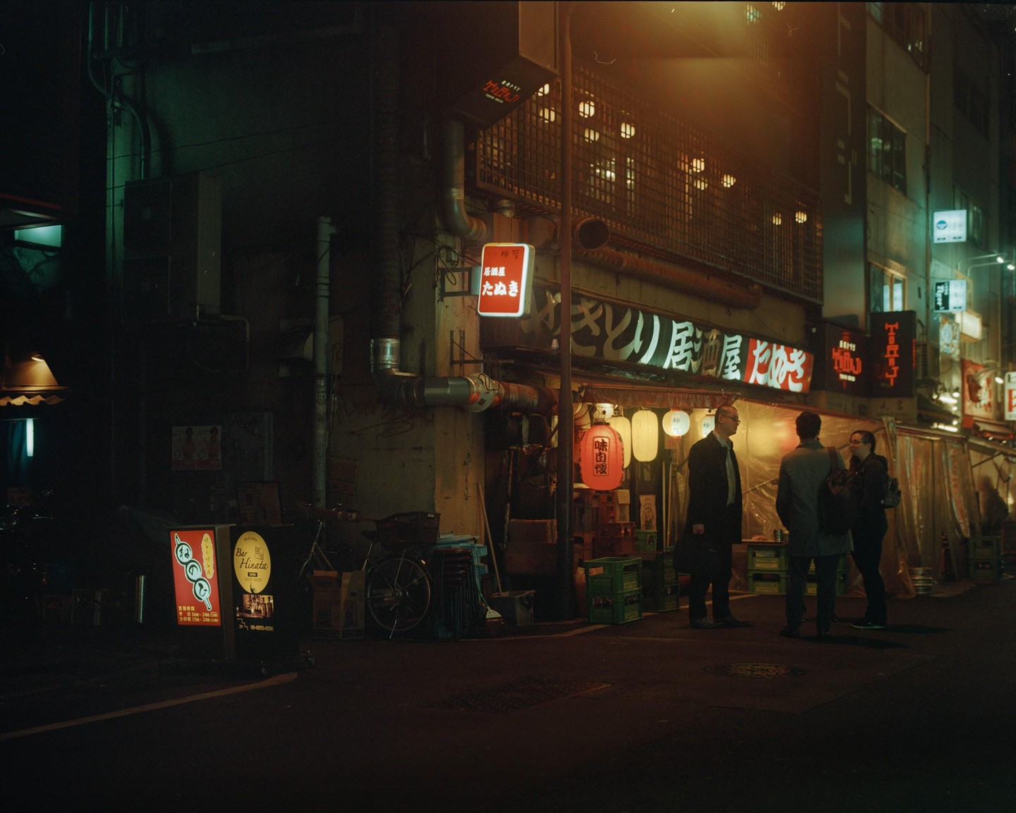 Three men stand on a street at night in front of a building with Japanese signs and lanterns.
