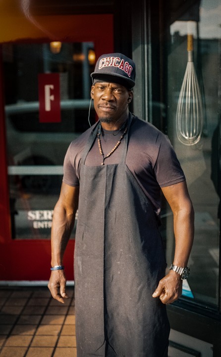 A man wearing an apron and a Chicago baseball cap stands in front of a storefront.
