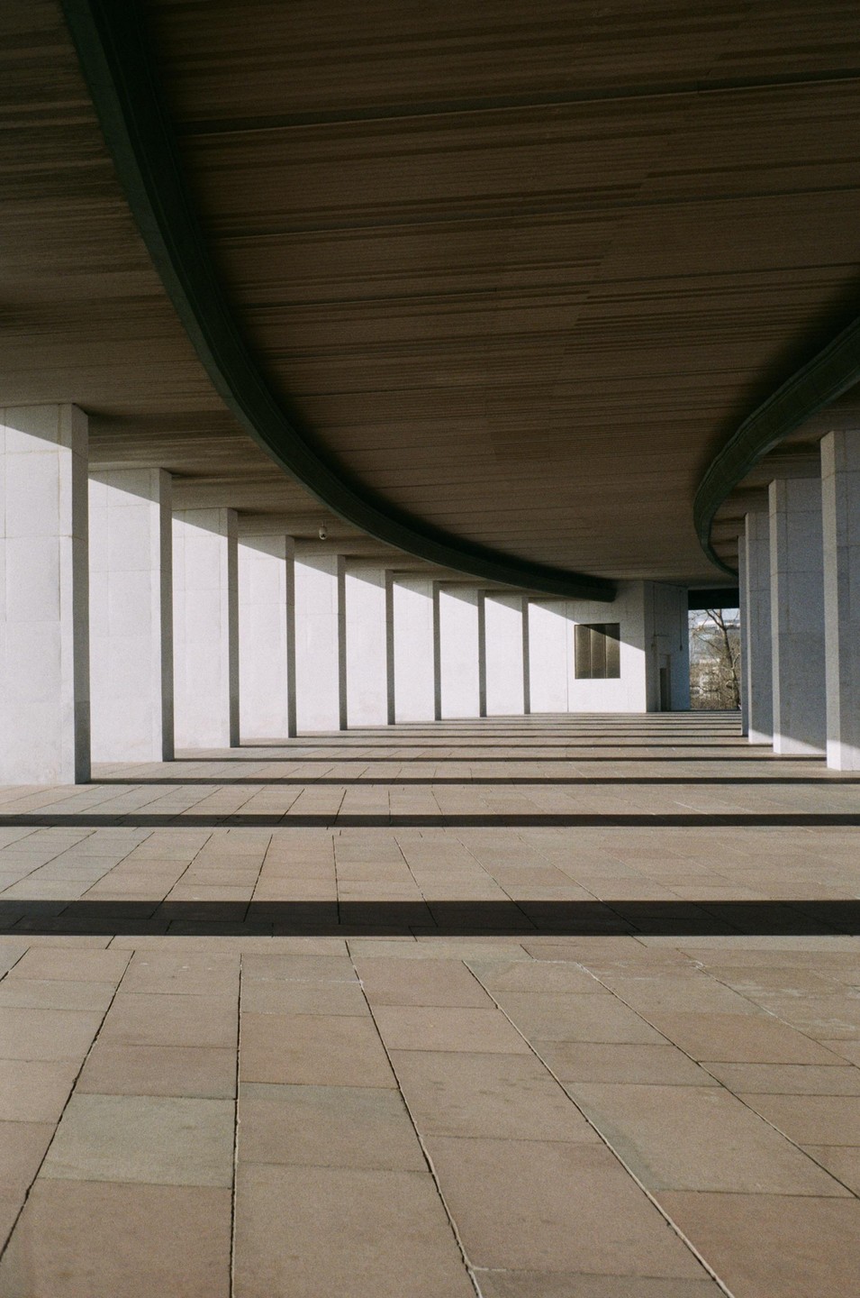 Sunlight casts long shadows across a tiled walkway lined with pillars under a curved concrete ceiling.