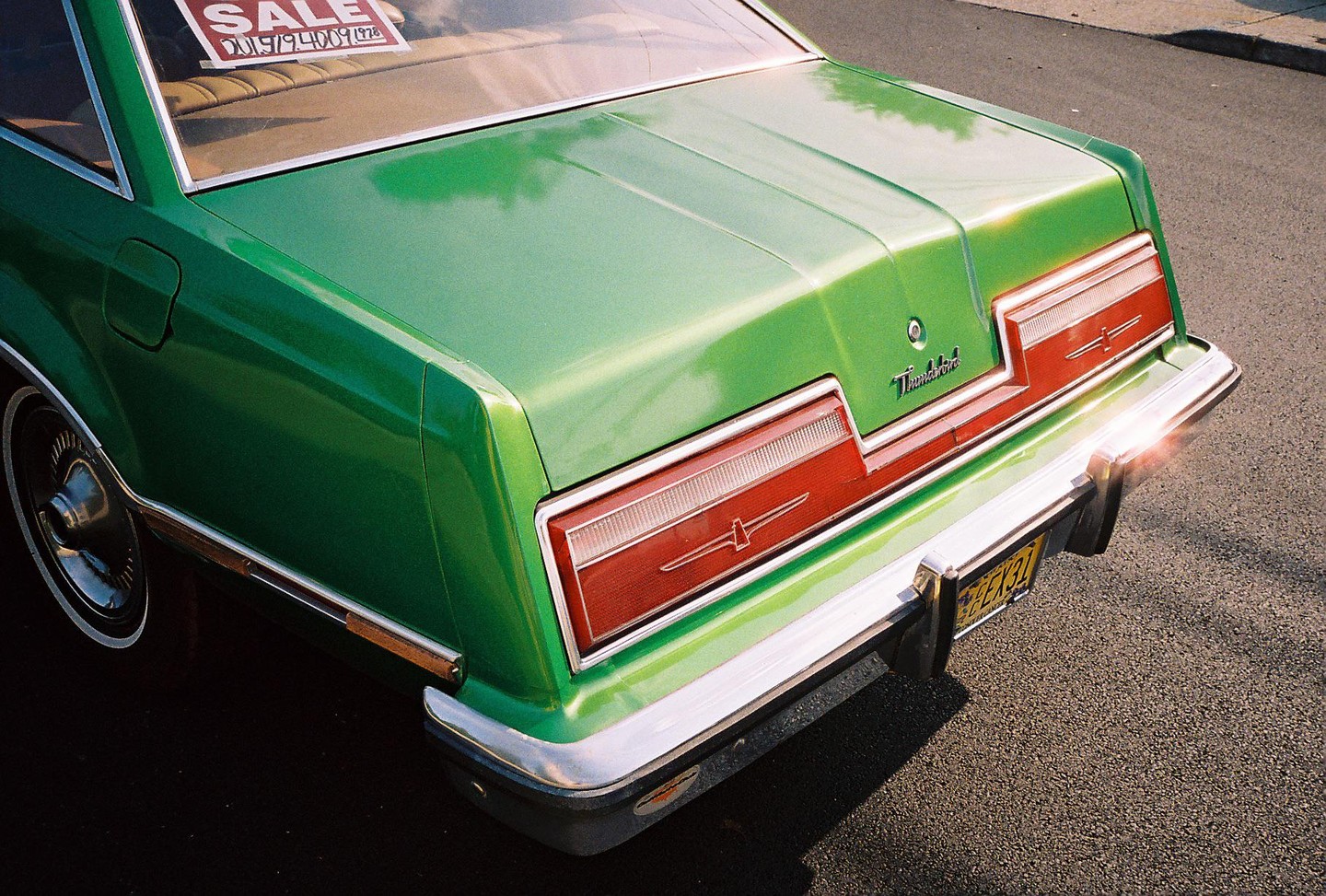 The rear of a green Ford Thunderbird with a "SALE" sign in the back window is parked on an asphalt street.