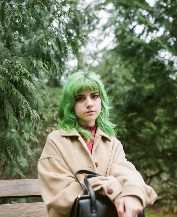 A woman with bright green hair and a beige coat sits on a wooden bench in front of green foliage.