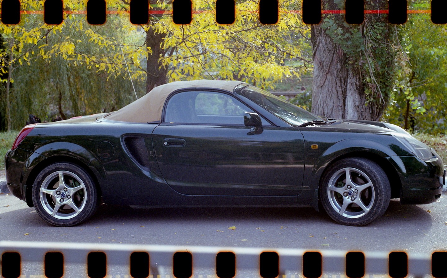A dark green convertible car with a tan soft top is parked on a paved surface next to a tree with yellow autumn foliage.