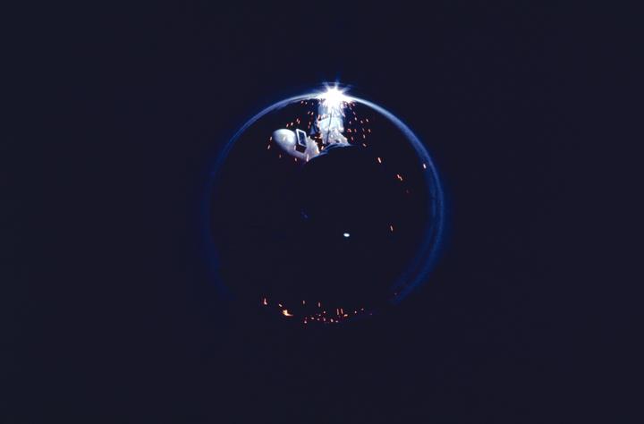 A welder works on a pipeline at night, surrounded by sparks and a circular light trail.