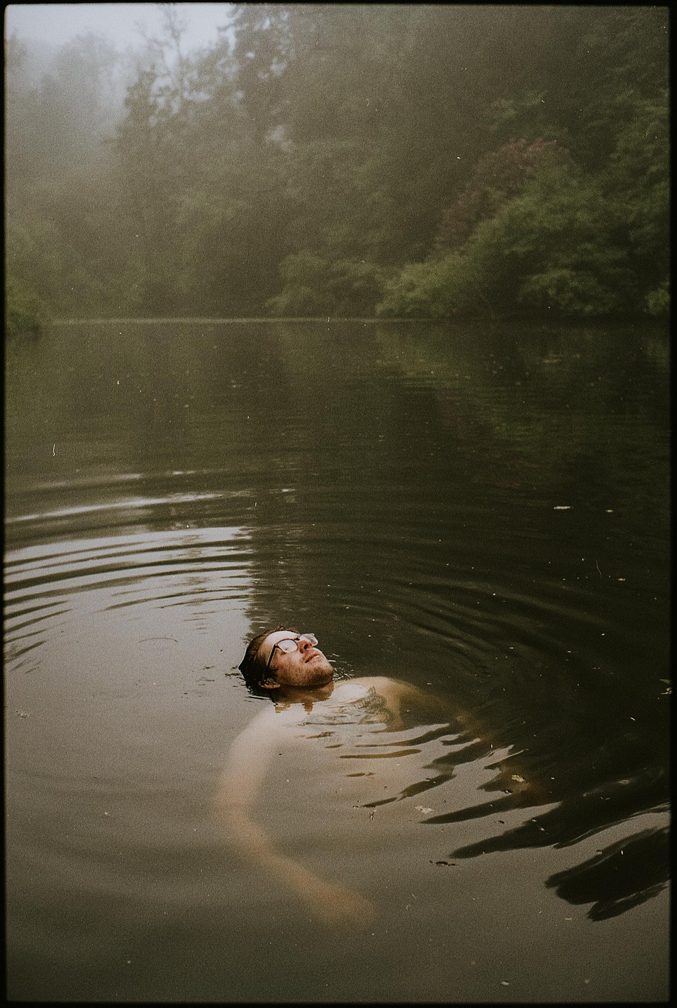 A man with glasses floats on his back in a lake surrounded by trees.