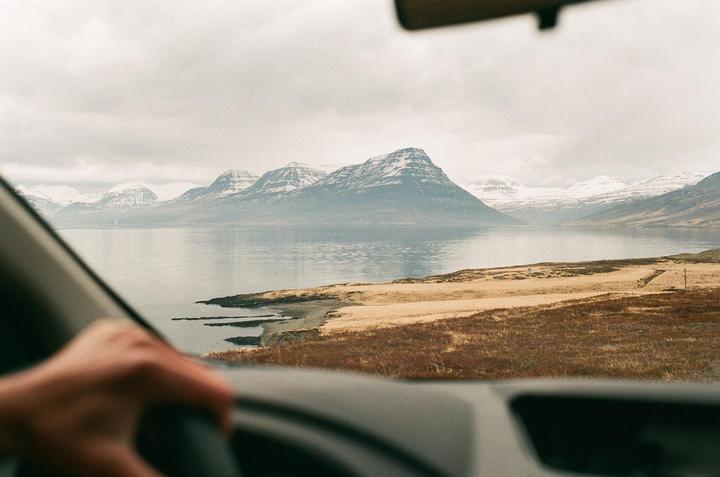 A hand is visible on a steering wheel, with a view of a lake and snow-capped mountains through the car windshield.