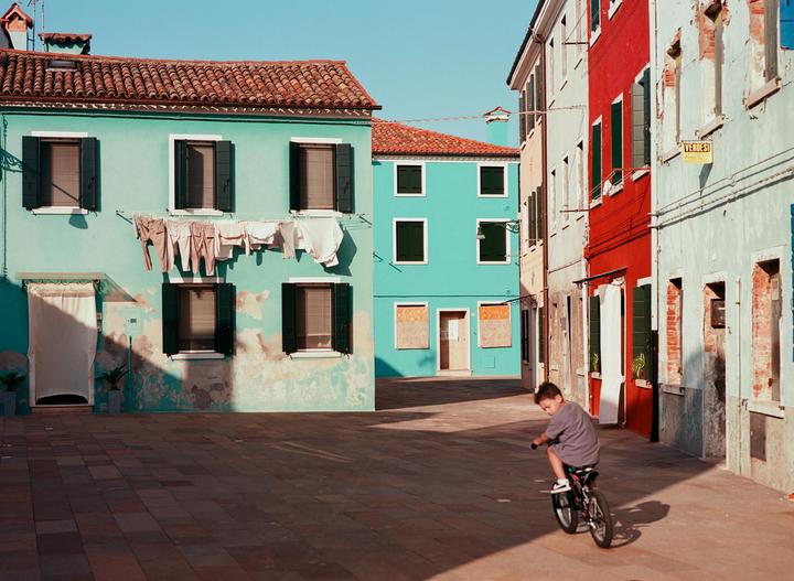 A boy rides a bicycle in a paved square surrounded by colorful buildings with laundry hanging from a line.
