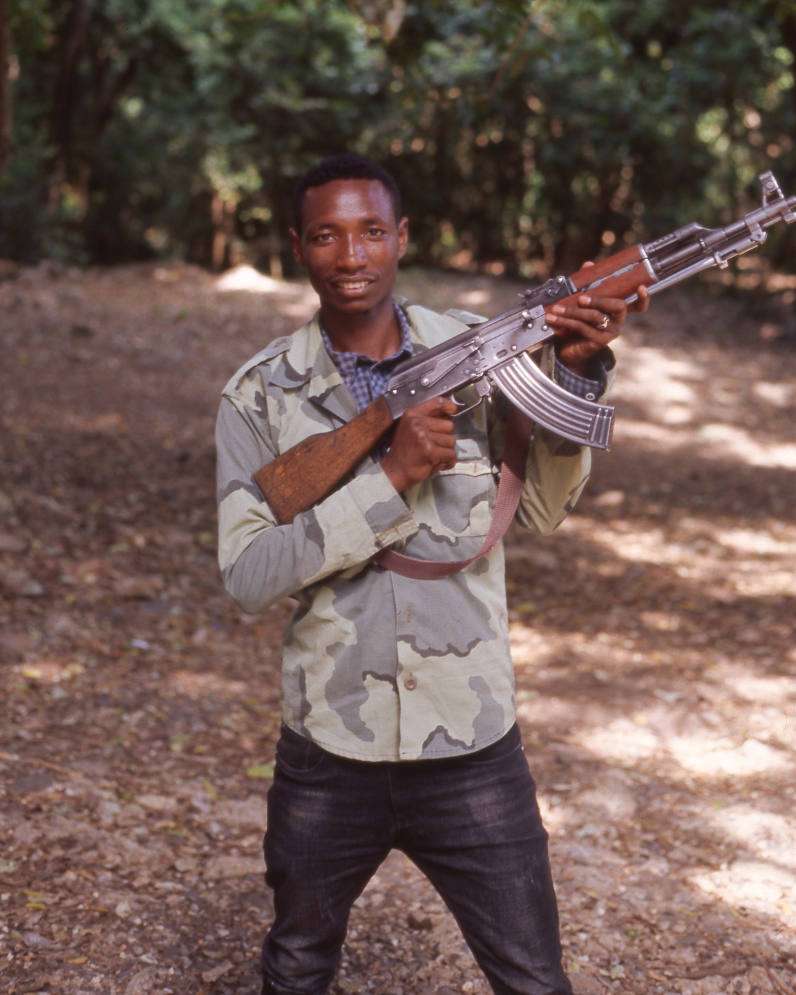 A man in a camouflage shirt holds a rifle in a forest.