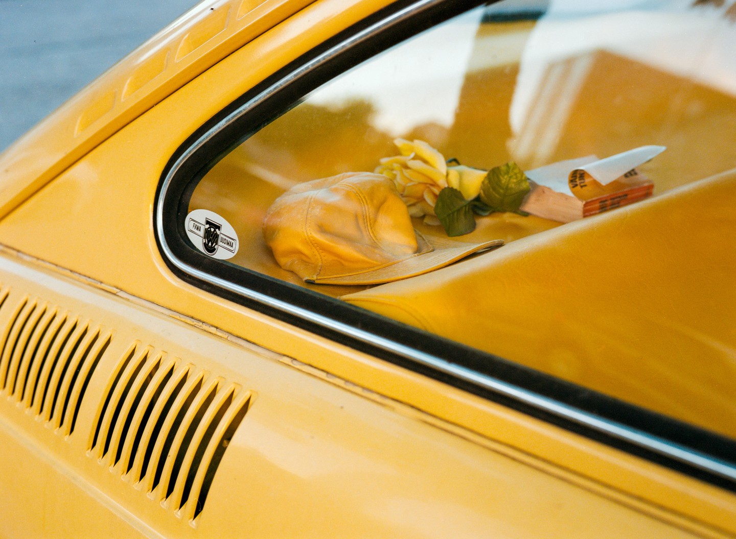 A yellow hat, a yellow rose, and a book are visible through the window of a yellow car.