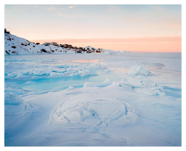 A frozen sea with ice formations and a distant village under a pink sky.