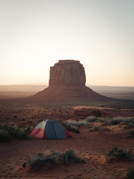 A red and gray tent is pitched in a desert landscape with a large mesa in the background.