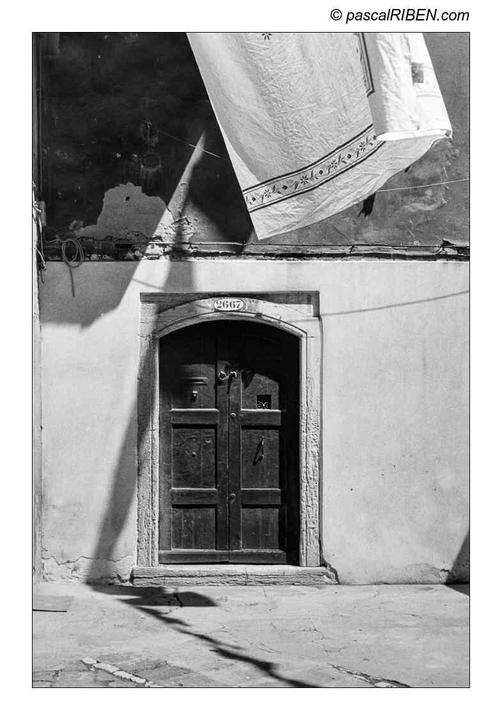 A dark wooden door with an arched frame is set into a light-colored wall, with a white sheet hanging above it and shadows cast on the ground.