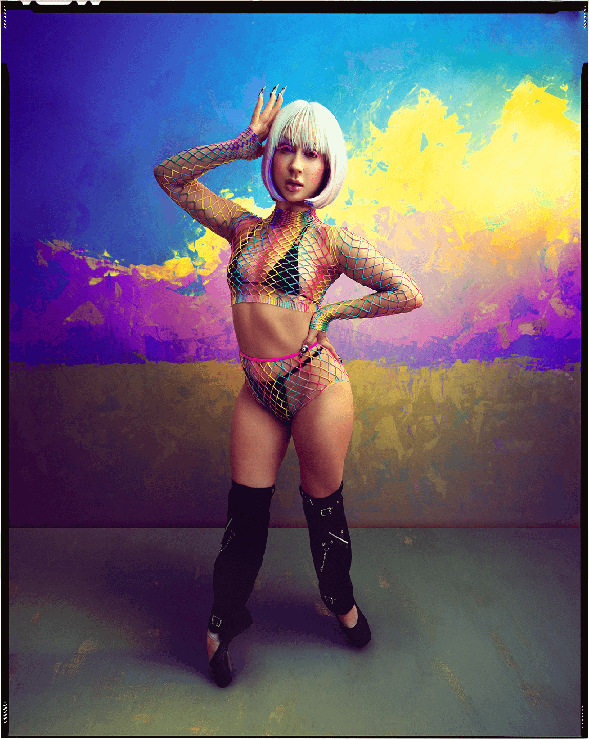 A woman in a colorful fishnet outfit and black boots poses against a vibrant painted backdrop.