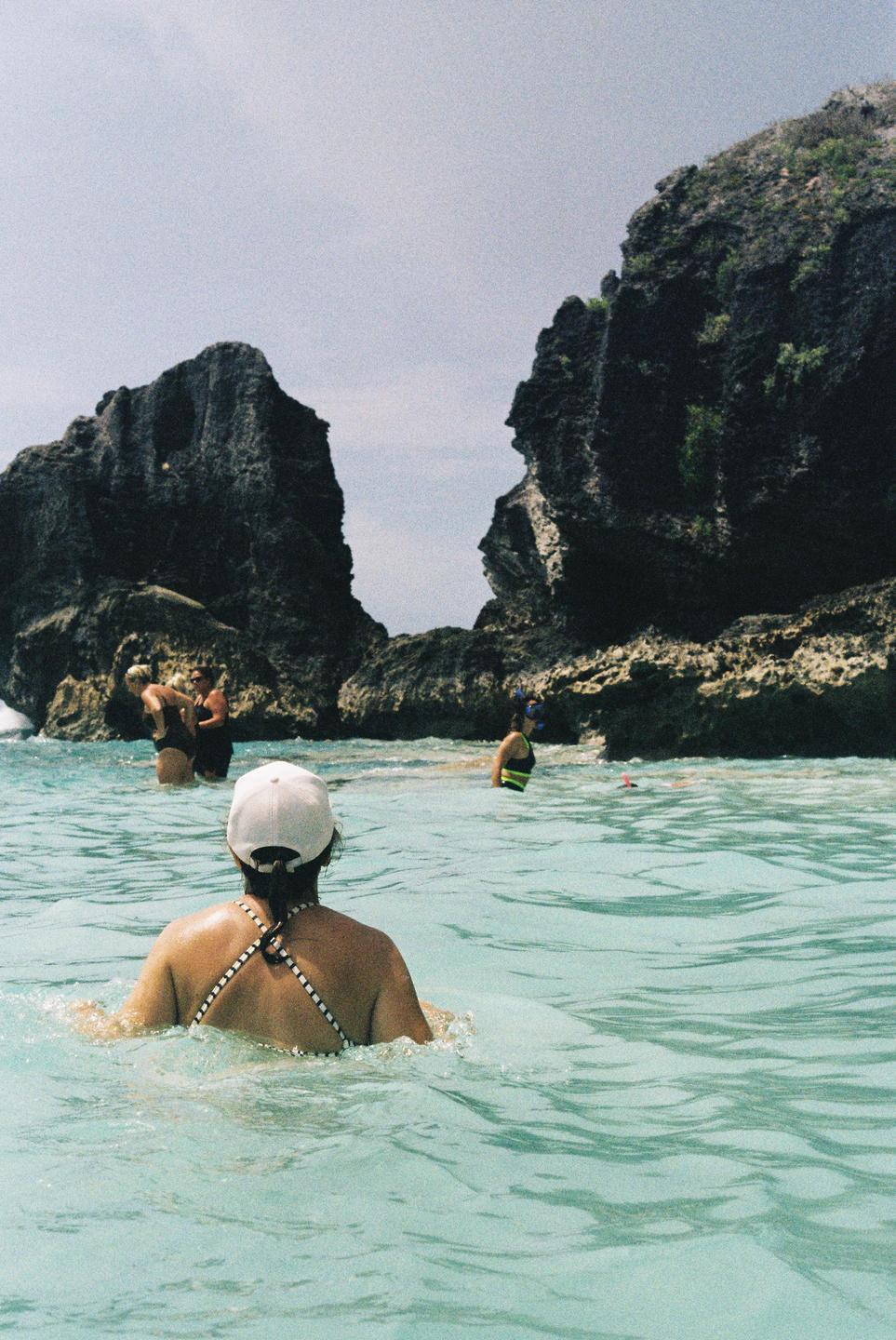 A woman in a white cap and striped bikini top is in the water with other people and large rock formations in the background.